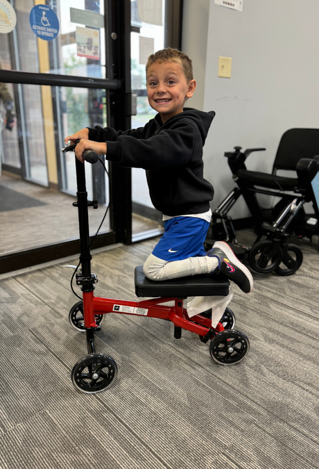 Smiling child riding a red Medline Junior Economy Knee Walker with black padded knee rest and handlebars, designed for safe mobility during leg injury recovery.