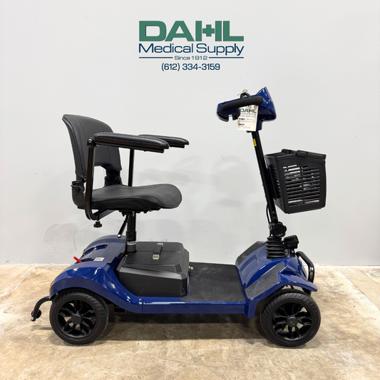 Blue used 4-wheel mobility scooter shown from the side, featuring a padded seat, armrests, front basket, and compact design, photographed inside Dahl Medical Supply.