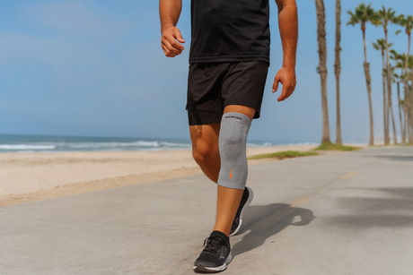 Man walking along a beach path wearing a gray knee brace for joint support and compression—comfortable knee support available at Dahl Medical Supply.