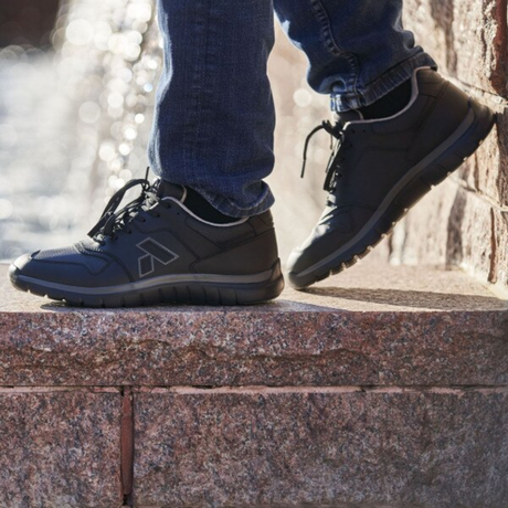 Person wearing black Anodyne No. 50 Sport Jogger shoes standing on a stone ledge in jeans, showcasing the shoes in an outdoor, lifestyle setting with a blurred water background.
