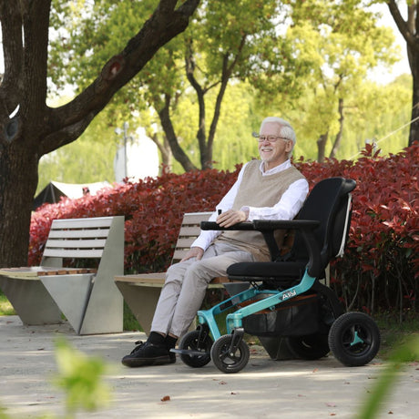 Adult seated comfortably in a Golden Ally GP303 portable power wheelchair outdoors, enjoying a park setting with trees, benches, and bright foliage in the background.