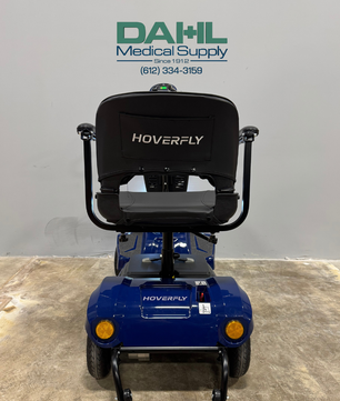 Rear view of a blue used 4-wheel mobility scooter showing the seat back, rear wheels, and stable frame, photographed inside a medical equipment showroom.