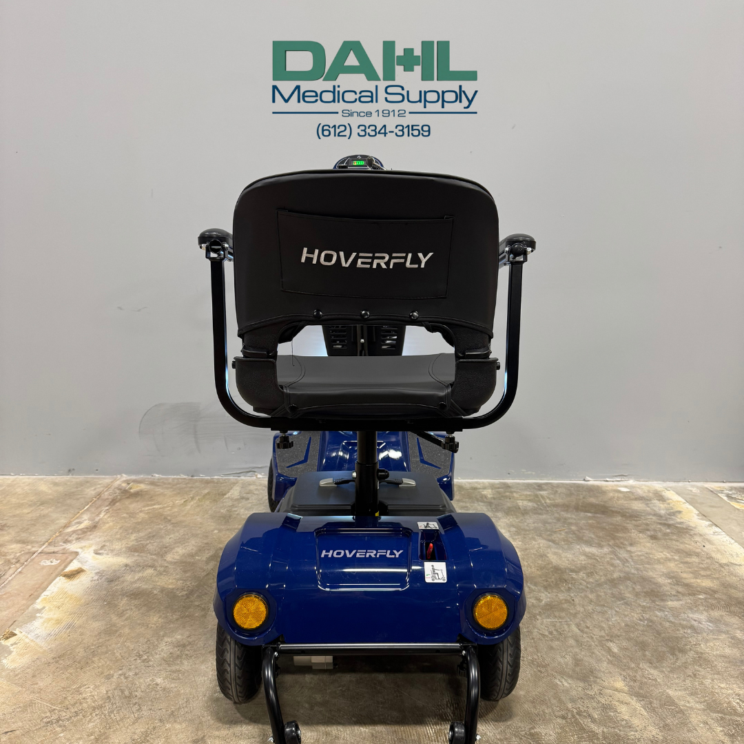 Rear view of a blue used 4-wheel mobility scooter showing the seat back, rear wheels, and stable frame, photographed inside a medical equipment showroom.