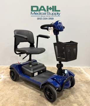 Angled front view of a blue used 4-wheel mobility scooter with front basket, padded seat, and black wheels, displayed indoors at Dahl Medical Supply.