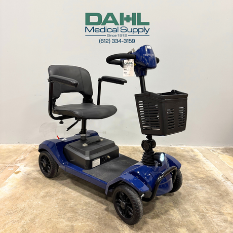 Angled front view of a blue used 4-wheel mobility scooter with front basket, padded seat, and black wheels, displayed indoors at Dahl Medical Supply.