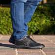 Close-up of a person wearing black Anodyne No. 50 Sport Jogger shoes with blue jeans, standing outdoors on a brick walkway with greenery in the background.