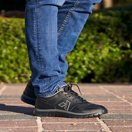 Close-up of a person wearing black Anodyne No. 50 Sport Jogger shoes with blue jeans, standing outdoors on a brick walkway with greenery in the background.